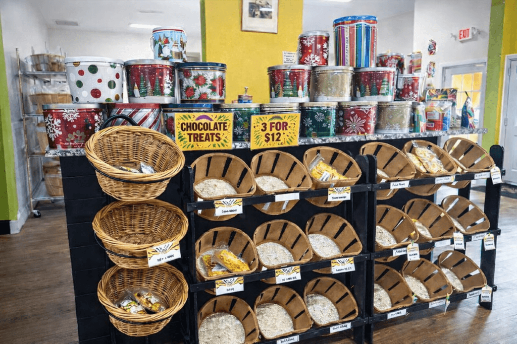 Interior of Wild About Popcorn shop with wooden popcorn bins, wicker baskets of treats, holiday-themed tins, and a chocolate treats promotion.