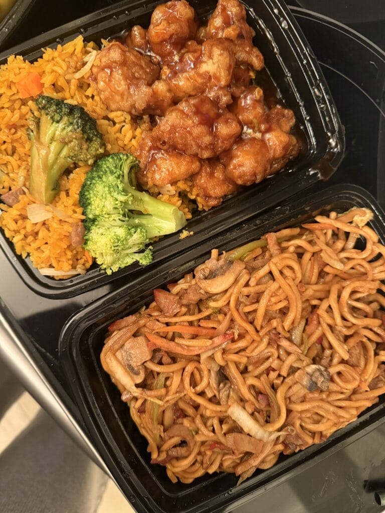 Takeout China Garden food in black containers with orange chicken over fried rice and broccoli, alongside lo mein noodles with vegetables.