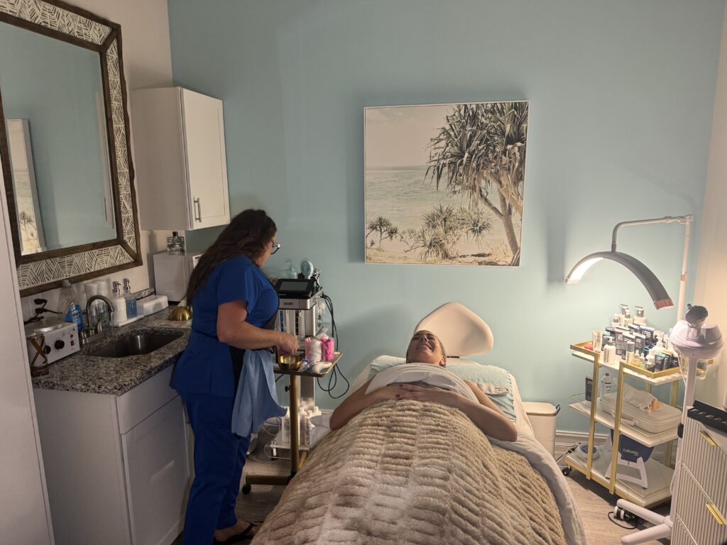 Esthetician at Glowing Results Skin Spa preparing facial treatment in a spa room while client relaxes on treatment bed.