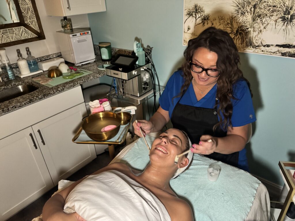 Esthetician at Glowing Results Skin Spa applying facial mask with brushes while client relaxes on treatment bed in spa room.