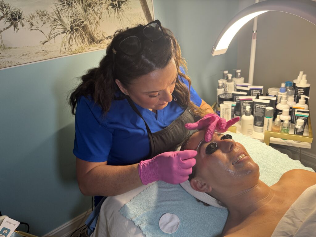 Esthetician at Glowing Results Skin Spa performing dermaplaning on client’s eyebrows while client relaxes on treatment bed in spa setting.