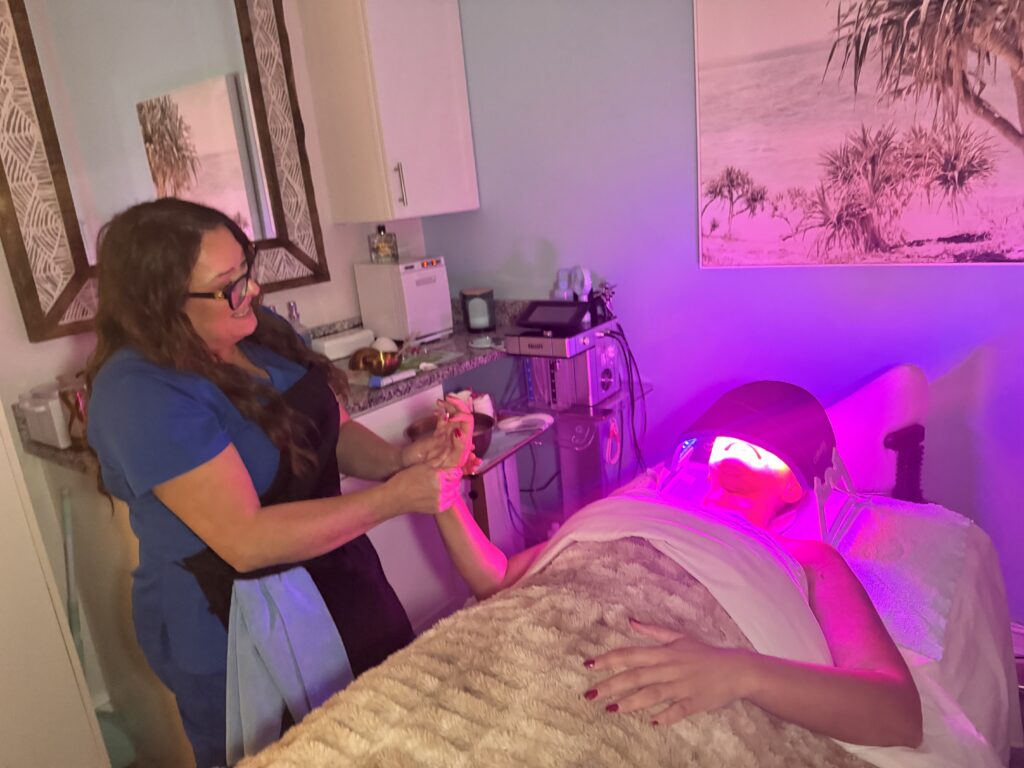 Client at Glowing Results Skin Spa receiving LED light therapy while esthetician gently massages her hand in spa setting.