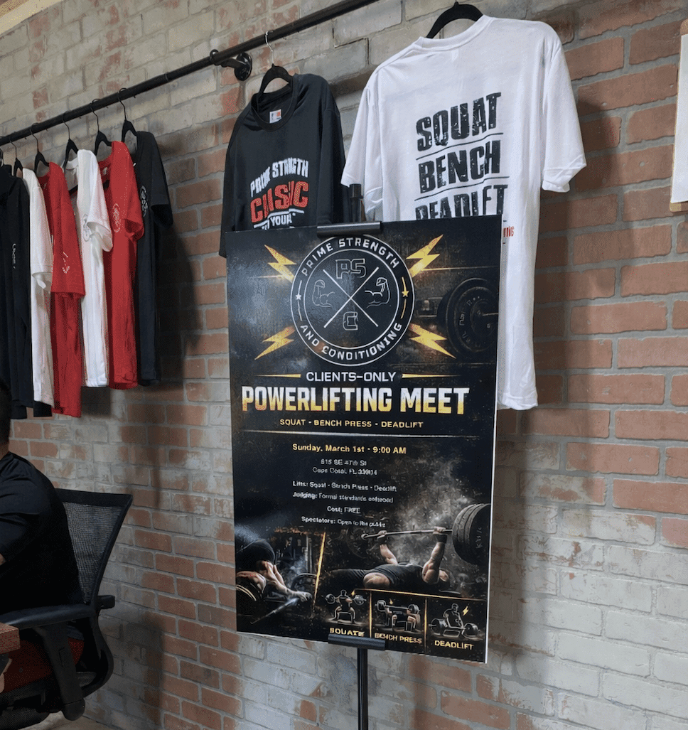 Powerlifting meet sign for Prime Strength and Conditioning in Cape Coral, FL, displayed inside a gym with a brick wall backdrop and branded apparel hanging nearby.
